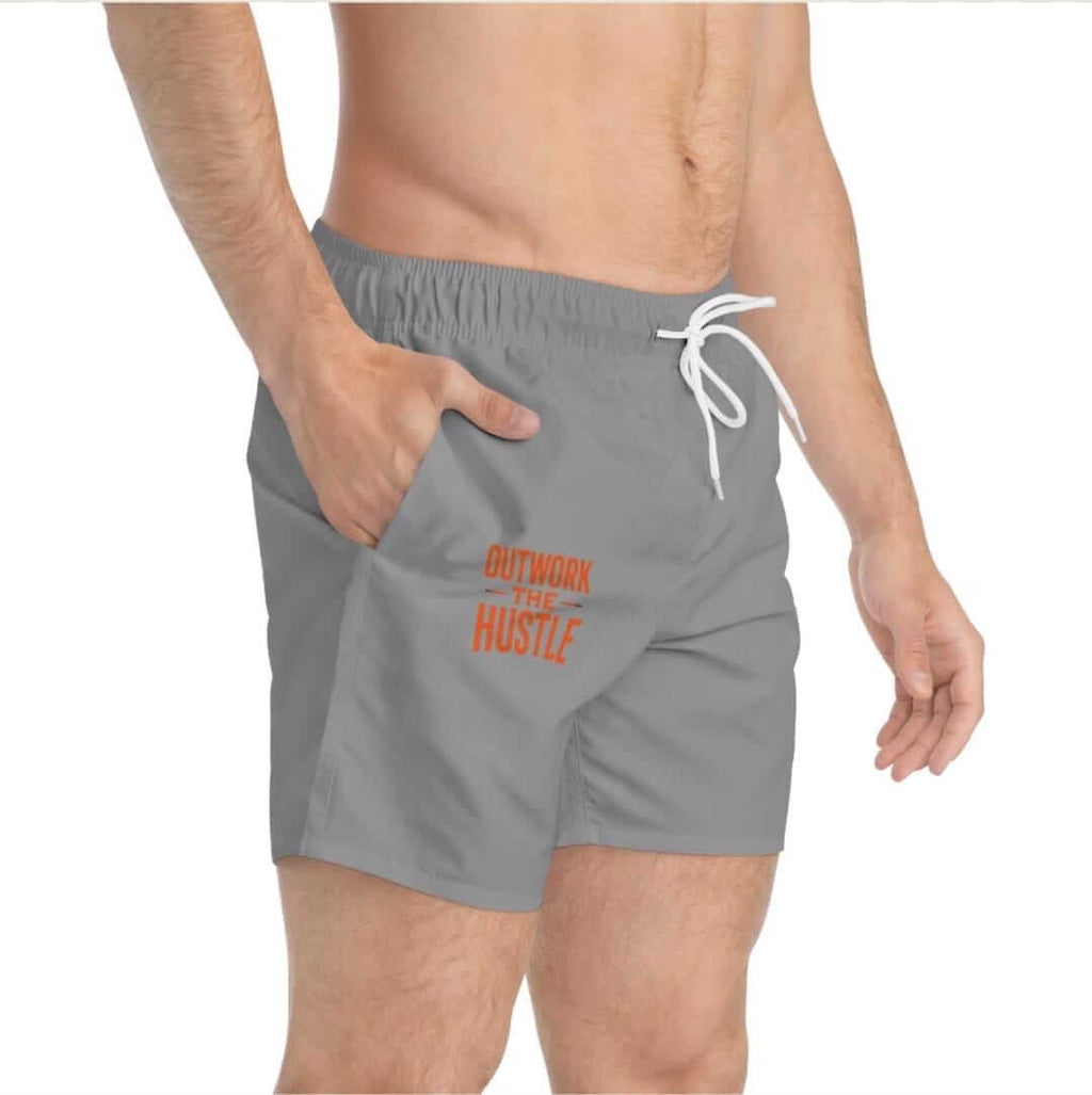 A model showcasing Outwork The Hustle Swim Trunks in gray with orange lettering.